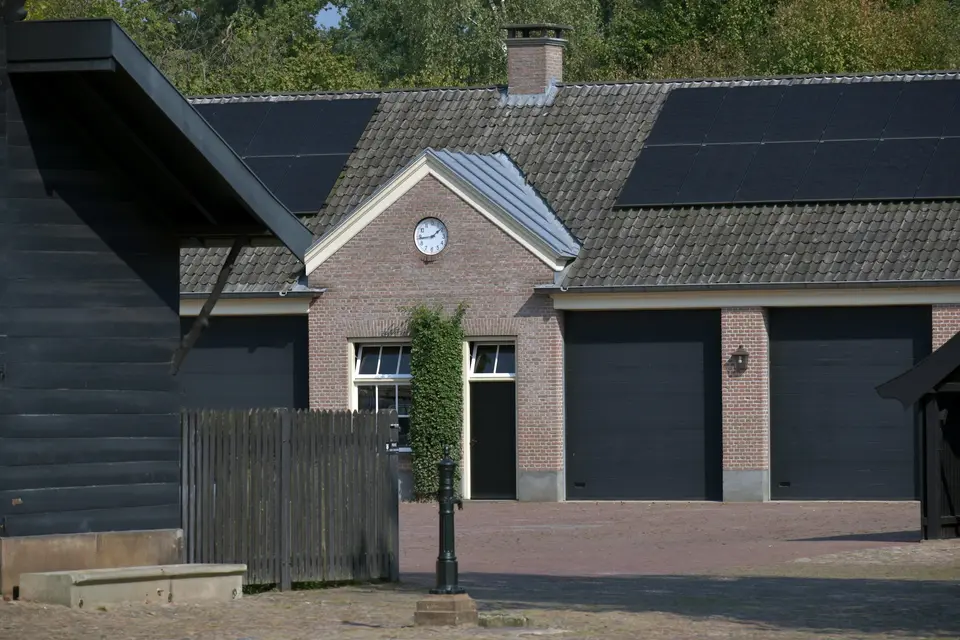 Een bakstenen koetshuis met een klok in de gevel en zwarte zonnepanelen op het pannendak, met een houten schutting en een pomp op het erf